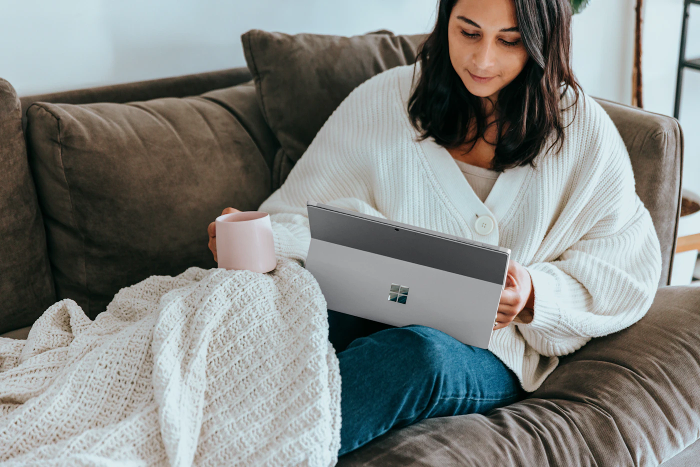 A woman relaxing on a couch under a knitted blanket with a warm drink, talking on her laptop