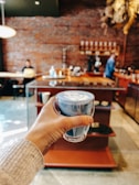 A hand holding a glass mug with blue-colored latte art, set against the backdrop of a cozy coffee shop. The interior features exposed brick walls, wooden shelves, and a counter with coffee-making equipment. In the background, a person is sitting with a laptop, while another person stands near the counter.