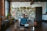 A person sits cross-legged in a cozy room with a laptop, two yoga blocks, and a lit candle nearby. The room features a wooden floor, a brick wall with a large window, and a wall of bookshelves adorned with cascading houseplants. Comfortable seating arrangements and potted plants add to the serene atmosphere.