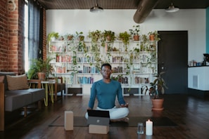 A person sits cross-legged in a cozy room with a laptop, two yoga blocks, and a lit candle nearby. The room features a wooden floor, a brick wall with a large window, and a wall of bookshelves adorned with cascading houseplants. Comfortable seating arrangements and potted plants add to the serene atmosphere.