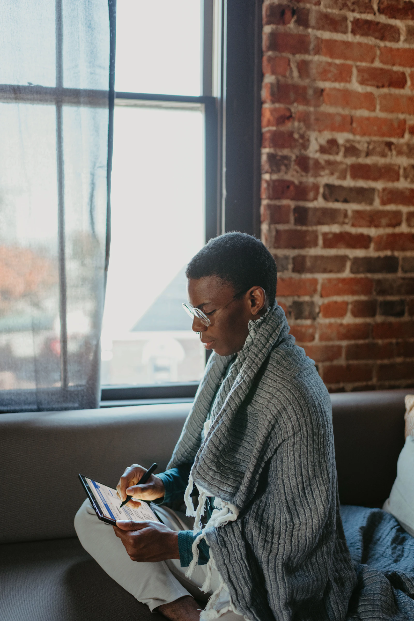 Student using a tablet with stylus while sitting on a couch near a window