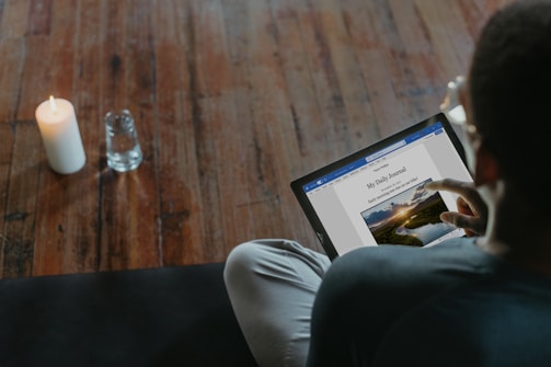 A person is seated on a dark surface, interacting with a laptop or tablet and reading a digital journal titled 'My Daily Journal'. A lit candle and a glass of water are placed on a wooden floor nearby.