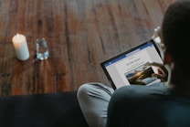 A person is seated on a dark surface, interacting with a laptop or tablet and reading a digital journal titled 'My Daily Journal'. A lit candle and a glass of water are placed on a wooden floor nearby.