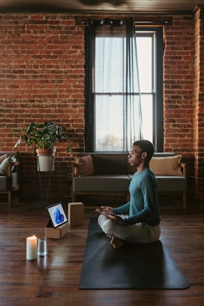 A person sits cross-legged on a yoga mat in a peaceful living room with exposed brick walls and wooden floors. Soft, natural light filters through a large window covered by sheer curtains. Nearby, a lit candle and a glass of water are placed on the floor beside a tablet displaying an abstract design. A green plant is seen next to a couch with cushions in the background.