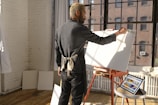 A person with an apron stands in a sunlit studio by a large window, working on a blank canvas on an easel. The floor is wooden, and there are tablets with colorful digital art resting on a nearby table. The walls are white and exposed brick.