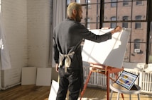 A person with an apron stands in a sunlit studio by a large window, working on a blank canvas on an easel. The floor is wooden, and there are tablets with colorful digital art resting on a nearby table. The walls are white and exposed brick.