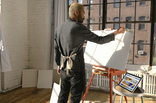 A person with an apron stands in a sunlit studio by a large window, working on a blank canvas on an easel. The floor is wooden, and there are tablets with colorful digital art resting on a nearby table. The walls are white and exposed brick.
