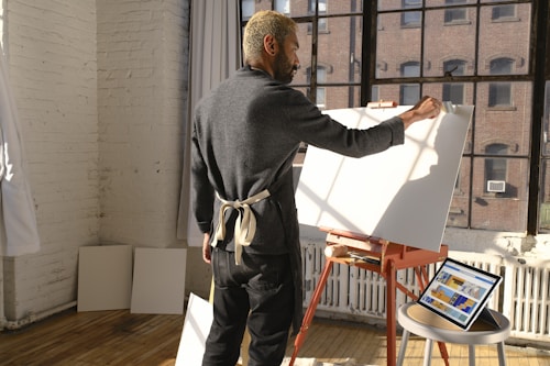 A person with an apron stands in a sunlit studio by a large window, working on a blank canvas on an easel. The floor is wooden, and there are tablets with colorful digital art resting on a nearby table. The walls are white and exposed brick.