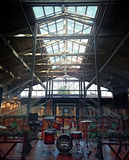 A spacious industrial interior with a high glass ceiling and metal beams. The area is well-lit with natural light streaming in. At the center, a drum set is positioned on a wooden stage, surrounded by greenery and decorative elements. Bricks and steel elements add to the rustic aesthetic.