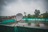 A rooftop with a satellite dish marked Tata Sky, set against a cloudy sky. The roof is bordered by a greenish-blue wall, and the wet surface suggests recent rain. In the background, there are trees and some brick buildings visible, giving a sense of an urban or semi-urban environment.