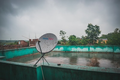 A rooftop with a satellite dish marked Tata Sky, set against a cloudy sky. The roof is bordered by a greenish-blue wall, and the wet surface suggests recent rain. In the background, there are trees and some brick buildings visible, giving a sense of an urban or semi-urban environment.