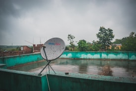 A rooftop with a satellite dish marked Tata Sky, set against a cloudy sky. The roof is bordered by a greenish-blue wall, and the wet surface suggests recent rain. In the background, there are trees and some brick buildings visible, giving a sense of an urban or semi-urban environment.