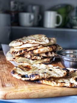 A stack of freshly cooked flatbread is placed on a wooden cutting board. The bread has a charred, golden-brown surface. In the background, there are several white and green coffee mugs on a shelf. A silver bowl is partially visible beside the bread.