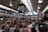 A bustling indoor market with a high, industrial-style ceiling filled with numerous people dining at communal tables. Overhead, there are large black and white signs indicating various food stalls. The photograph captures a busy and vibrant atmosphere.