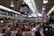 A bustling indoor market with a high, industrial-style ceiling filled with numerous people dining at communal tables. Overhead, there are large black and white signs indicating various food stalls. The photograph captures a busy and vibrant atmosphere.