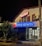 A building at night with a prominent blue neon sign that reads 'APPLE ESTATES'. Above, there is another sign in red that also includes 'APPLE' and additional contact details. The facade has wooden shutters and an air conditioning unit. Trees in planters are positioned near the entrance.