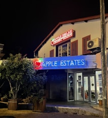 A building at night with a prominent blue neon sign that reads 'APPLE ESTATES'. Above, there is another sign in red that also includes 'APPLE' and additional contact details. The facade has wooden shutters and an air conditioning unit. Trees in planters are positioned near the entrance.