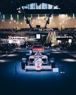 A vintage Formula 1 race car is displayed prominently under bright lights in an indoor exhibition setting. The surroundings include other classic vehicles and a decorated Christmas tree, adding a festive atmosphere. The ceiling has large, arched windows, and modern lighting fixtures accentuate the display.