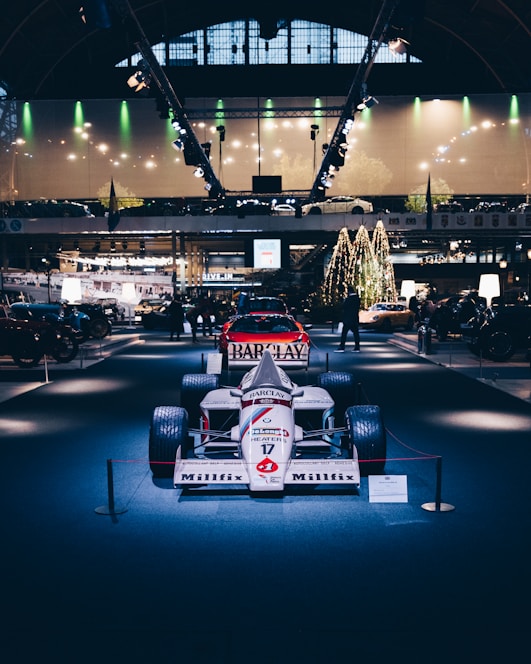 A vintage Formula 1 race car is displayed prominently under bright lights in an indoor exhibition setting. The surroundings include other classic vehicles and a decorated Christmas tree, adding a festive atmosphere. The ceiling has large, arched windows, and modern lighting fixtures accentuate the display.