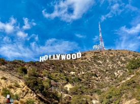 Large white letters spell out 'Hollywood' across a dry, vegetation-covered hillside under a bright blue sky. A tall communication tower stands nearby, surrounded by satellite dishes.