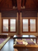A traditional room with wooden interiors featuring two large windows with wooden blinds that allow sunlight to filter through. A low table is placed on a polished wooden floor, with floor cushions and a floor mattress on the side, adorned with decorative patterns.