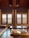 A traditional room with wooden interiors featuring two large windows with wooden blinds that allow sunlight to filter through. A low table is placed on a polished wooden floor, with floor cushions and a floor mattress on the side, adorned with decorative patterns.