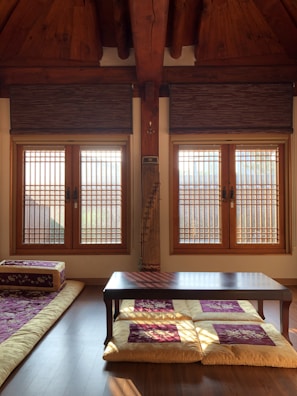 A traditional room with wooden interiors featuring two large windows with wooden blinds that allow sunlight to filter through. A low table is placed on a polished wooden floor, with floor cushions and a floor mattress on the side, adorned with decorative patterns.