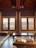 A traditional room with wooden interiors featuring two large windows with wooden blinds that allow sunlight to filter through. A low table is placed on a polished wooden floor, with floor cushions and a floor mattress on the side, adorned with decorative patterns.