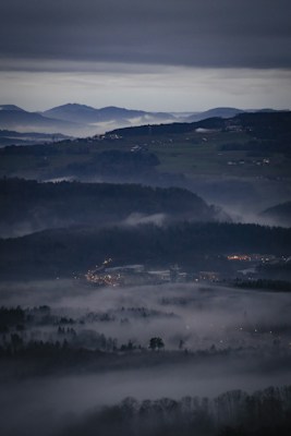 A vast, misty landscape features rolling hills and a small town with illuminated streetlights nestled among the fog. The horizon is marked by a range of distant mountains under an overcast sky, creating a serene, mysterious ambience.
