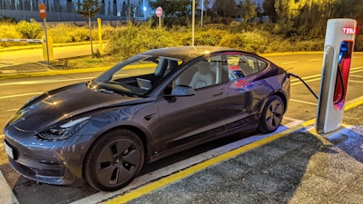 A sleek, modern electric car is parked at a charging station during twilight. The car is plugged into a Tesla Supercharger, surrounded by a dimly lit street with some vegetation in the background.