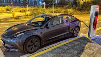 A sleek, modern electric car is parked at a charging station during twilight. The car is plugged into a Tesla Supercharger, surrounded by a dimly lit street with some vegetation in the background.