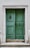 A pair of weathered green wooden doors set in a stone frame with a slightly worn appearance. The doors have metal accents, including a door knocker on the left side and a handle on the right. They are elevated by a small set of stone steps, and the surrounding wall is light in color, complementing the overall aged look.