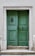 A pair of weathered green wooden doors set in a stone frame with a slightly worn appearance. The doors have metal accents, including a door knocker on the left side and a handle on the right. They are elevated by a small set of stone steps, and the surrounding wall is light in color, complementing the overall aged look.