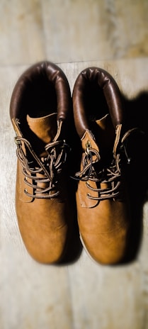 A pair of brown leather boots with dark brown padded collars is placed side by side on a light wooden surface. The boots have a sturdy appearance with thick laces and metal eyelets.
