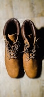 A pair of brown leather boots with dark brown padded collars is placed side by side on a light wooden surface. The boots have a sturdy appearance with thick laces and metal eyelets.