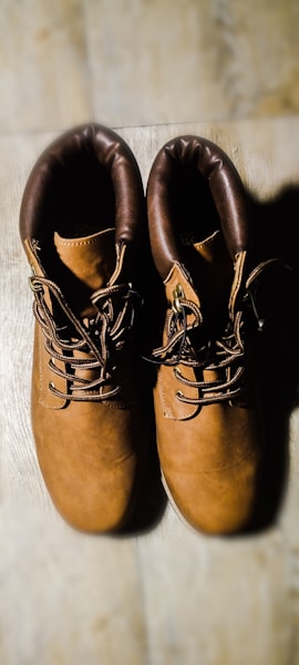A pair of brown leather boots with dark brown padded collars is placed side by side on a light wooden surface. The boots have a sturdy appearance with thick laces and metal eyelets.
