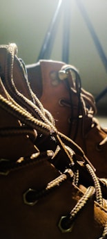 A close-up view of rugged brown leather boots with detailed laces and metal eyelets, set against a blurred background with warm lighting.