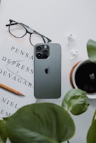 A neatly arranged flat lay featuring a smartphone, a pair of glasses, a book titled 'Pensar Depress e Devagar', a pencil, a pair of wireless earbuds, a cup of black coffee, and green leaves. The items are arranged on a white surface, creating a minimalist and organized aesthetic.
