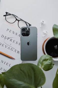 A neatly arranged flat lay featuring a smartphone, a pair of glasses, a book titled 'Pensar Depress e Devagar', a pencil, a pair of wireless earbuds, a cup of black coffee, and green leaves. The items are arranged on a white surface, creating a minimalist and organized aesthetic.