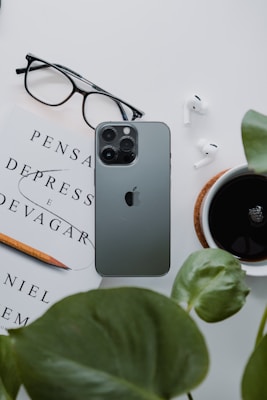 A neatly arranged flat lay featuring a smartphone, a pair of glasses, a book titled 'Pensar Depress e Devagar', a pencil, a pair of wireless earbuds, a cup of black coffee, and green leaves. The items are arranged on a white surface, creating a minimalist and organized aesthetic.