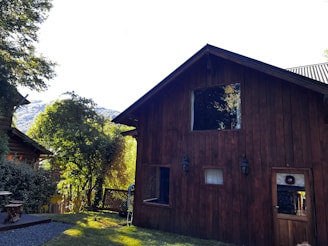 A rustic wooden house surrounded by lush greenery, with another wooden structure partially visible to the left. The architecture features dark wood paneling and a triangular roof. Sunlight highlights the foliage and creates a warm atmosphere. Mountains are visible in the background.
