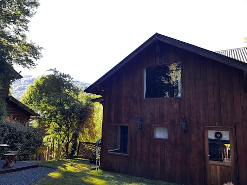 A rustic wooden house surrounded by lush greenery, with another wooden structure partially visible to the left. The architecture features dark wood paneling and a triangular roof. Sunlight highlights the foliage and creates a warm atmosphere. Mountains are visible in the background.