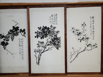 Three framed artworks feature traditional Chinese ink paintings depicting trees or plants with calligraphy accompanying each piece. The brushwork is expressive, with varying shades of black and gray creating depth and texture on the pale background.
