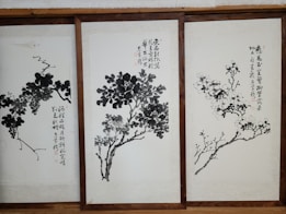 Three framed artworks feature traditional Chinese ink paintings depicting trees or plants with calligraphy accompanying each piece. The brushwork is expressive, with varying shades of black and gray creating depth and texture on the pale background.