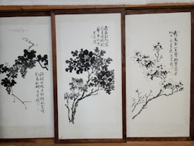 Three framed artworks feature traditional Chinese ink paintings depicting trees or plants with calligraphy accompanying each piece. The brushwork is expressive, with varying shades of black and gray creating depth and texture on the pale background.