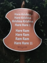 A sign with an orange background displays the Hare Krishna mantra in white text, surrounded by greenery and leaves.