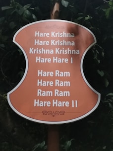 A sign with an orange background displays the Hare Krishna mantra in white text, surrounded by greenery and leaves.