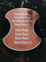 A sign with an orange background displays the Hare Krishna mantra in white text, surrounded by greenery and leaves.