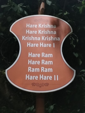 A sign with an orange background displays the Hare Krishna mantra in white text, surrounded by greenery and leaves.