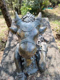 A bronze statue of a cow, intricately adorned with detailed decorations including beads and flower patterns, placed on a stone pedestal. The cow has its eyes closed and appears calm, with a backdrop of a wooded area with trees and some greenery.